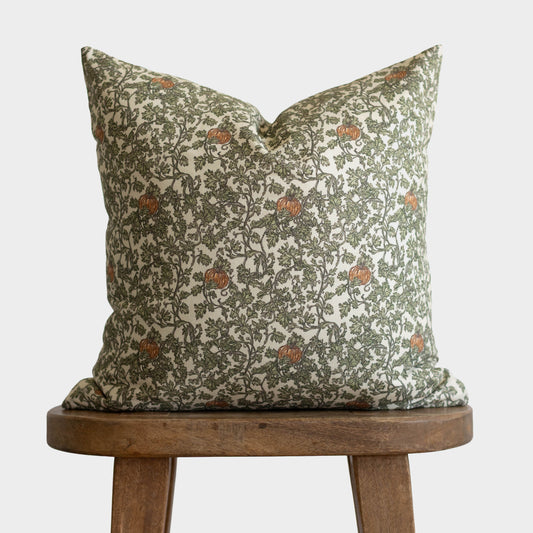 Cream linen pillow with olive green foliage and rust-colored gourds pattern displayed on wooden stool.