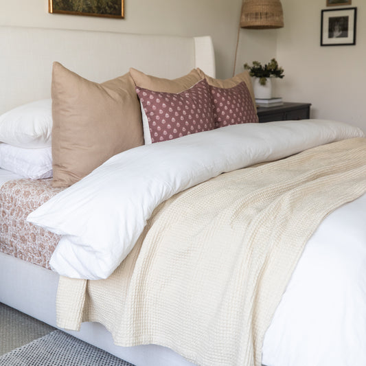 Ivory waffle-weave blanket draped across a bed with white pillows and decorative cushions.