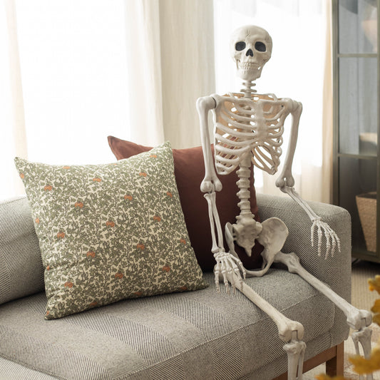 Cream-colored skeleton sitting on gray upholstered chair with green patterned pillow and burgundy cushion.