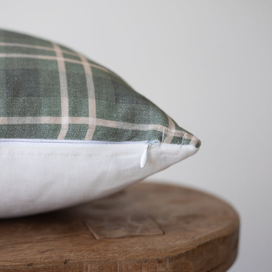 Green and white plaid pillow with white zipper detail resting on wooden round surface.
