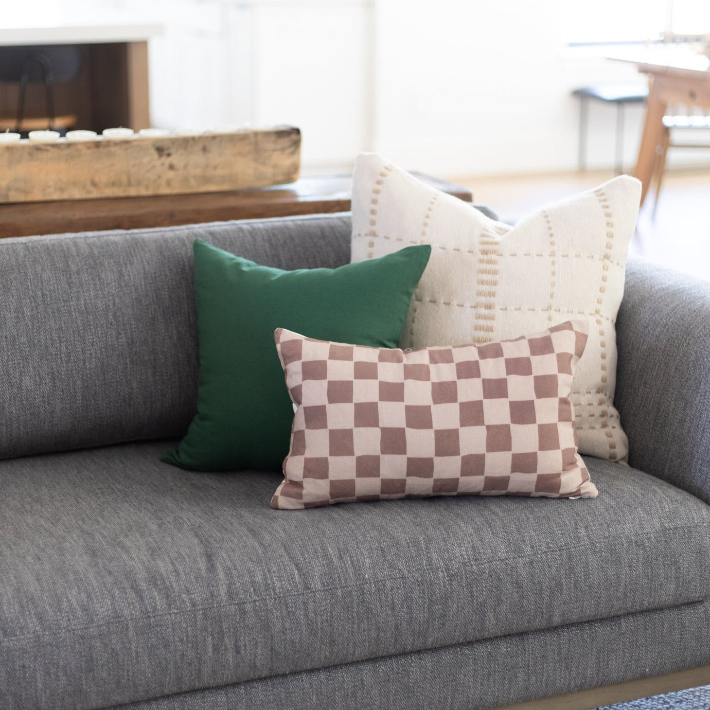 Three throw pillows on a gray sofa: solid green, checkerboard pink and white, and cream with geometric pattern.
