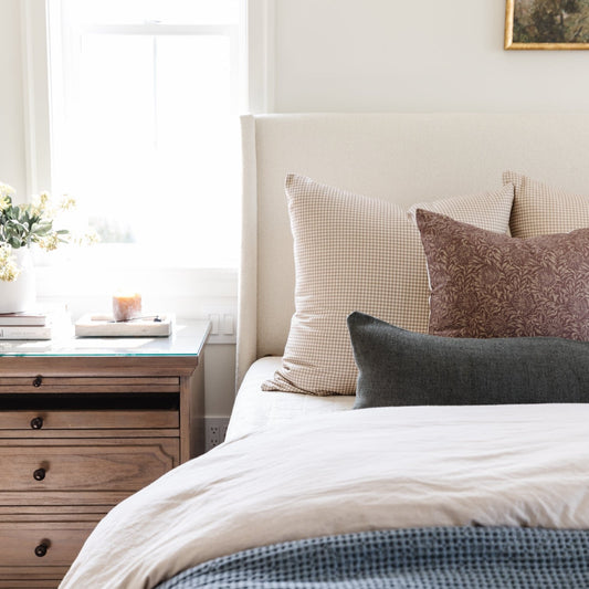 Tan and white gingham patterned pillow on a bed with neutral bedding and wooden nightstand.