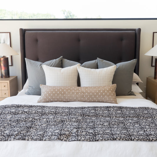 Beige lumbar pillow with white polka dot pattern displayed on bed between grid-patterned pillows.