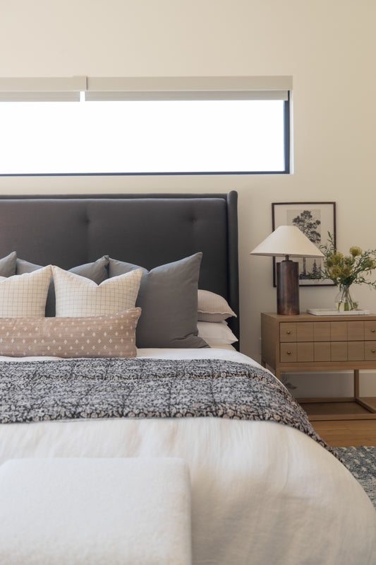 Bedroom with dark gray upholstered headboard, layered pillows in neutral tones, textured bedding, wooden nightstand with lamp