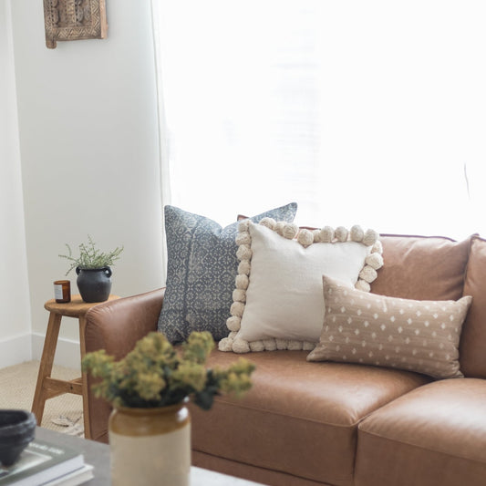 Cream lumbar pillow with pom-pom trim displayed on brown leather sofa with patterned pillows.