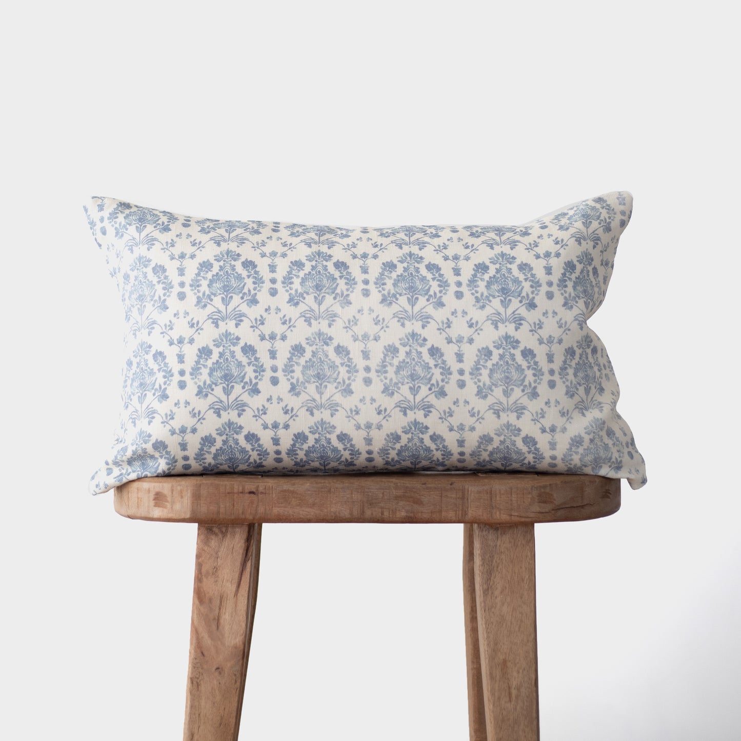 Blue and white damask patterned linen pillow on wooden bench against white background.