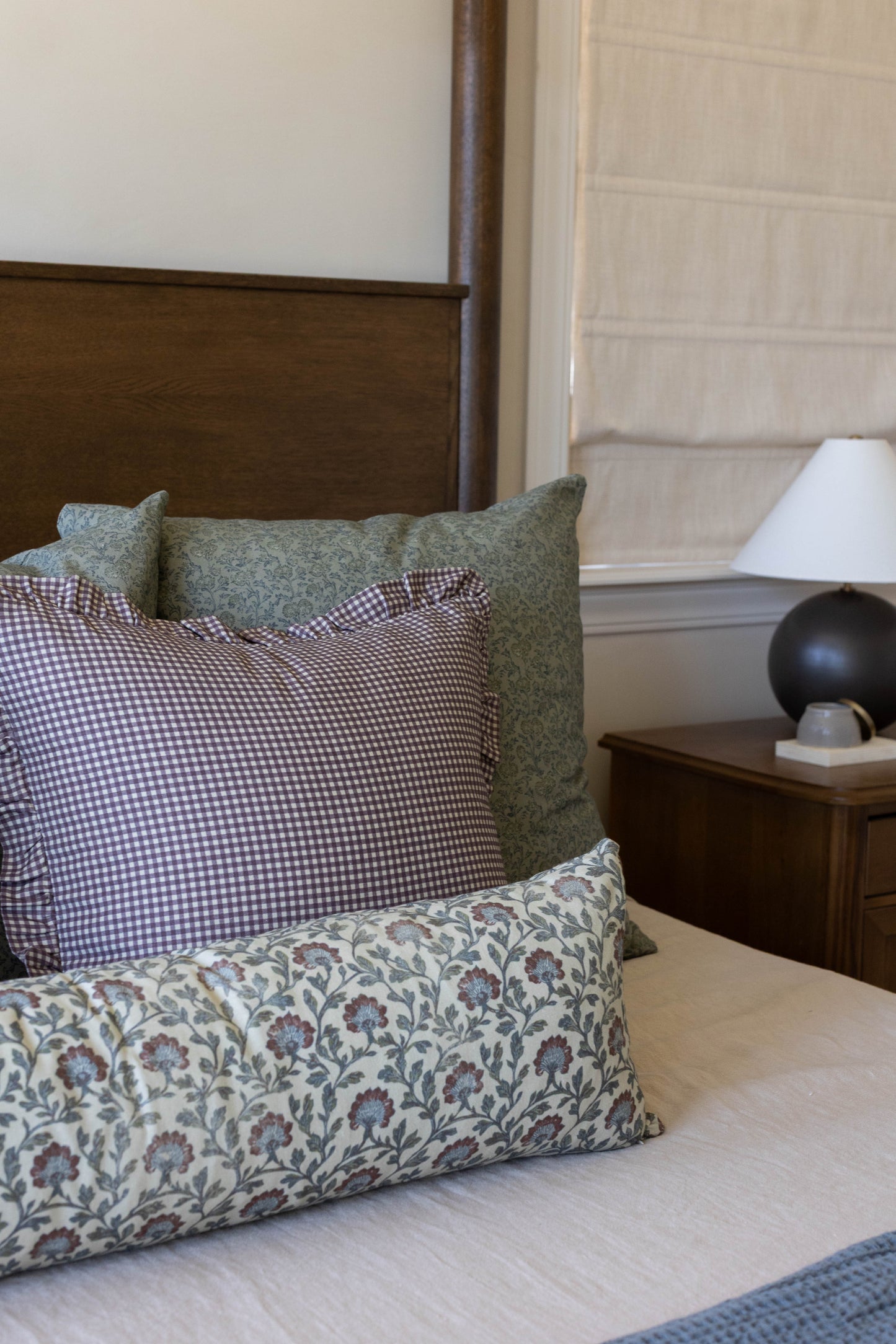 Plum and white gingham ruffle pillow displayed on bed with green and floral patterned pillows.