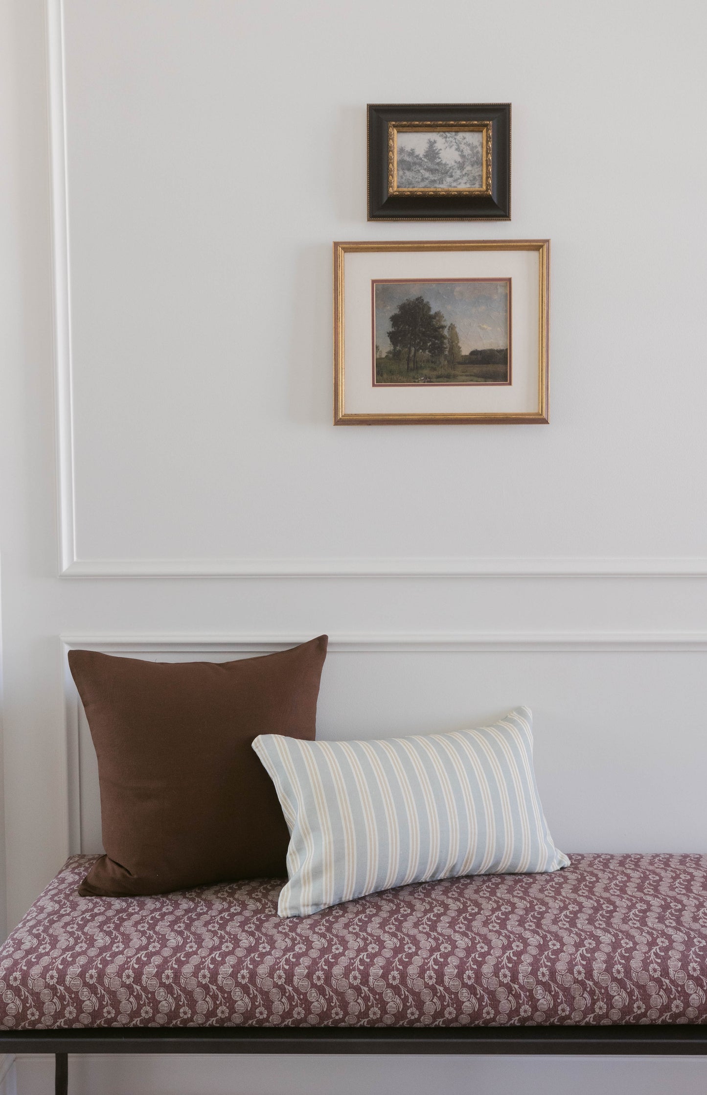 Bedroom bench with burgundy patterned cushion, brown and blue striped pillows, and two framed landscape artworks on wall abov