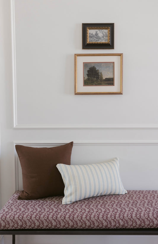 Brown square pillow and blue striped rectangular pillow on patterned bedding against white wall with two framed landscape art