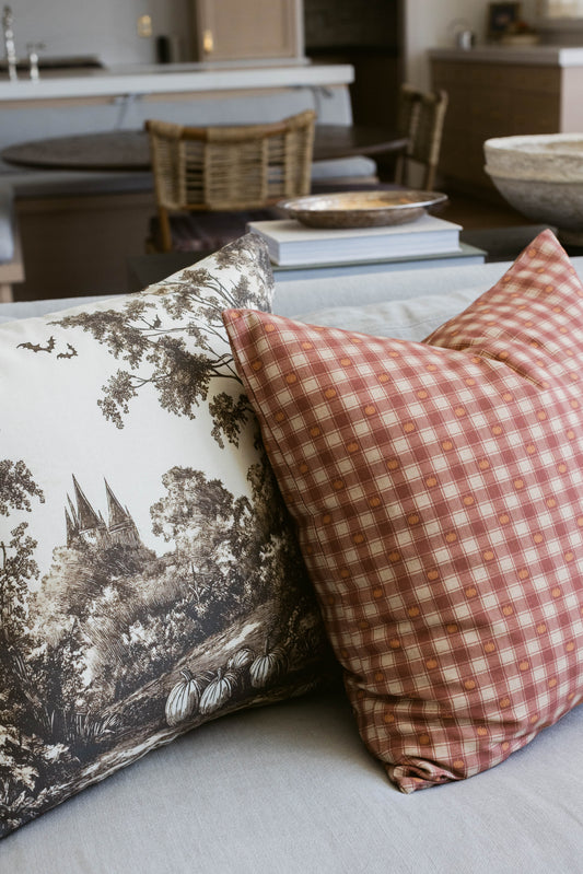 Two decorative pillows on a table: one with sepia-toned forest and castle illustration, one with coral and white gingham chec
