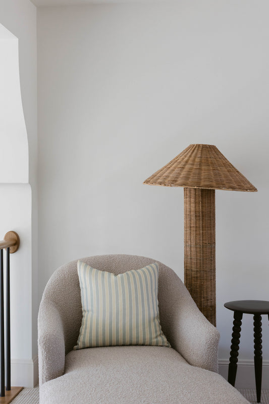Woven rattan floor lamp with conical shade and cylindrical base next to gray upholstered armchair.