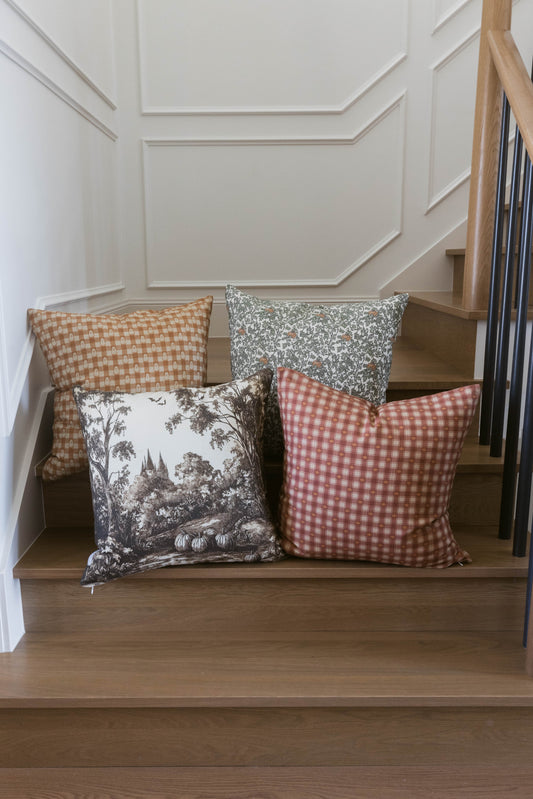 Four decorative pillows with checkered and botanical patterns in tan, gray, and red displayed on wooden stairs.