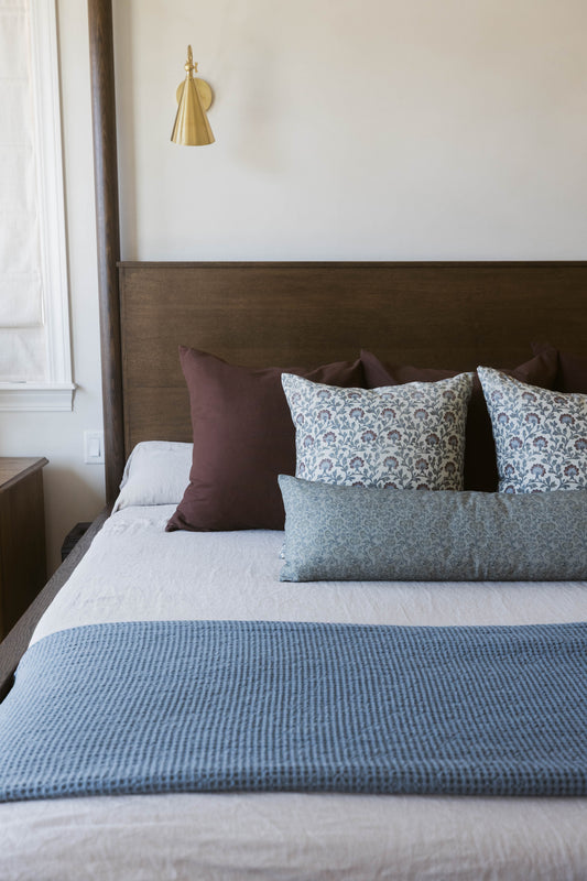 Brass cone pendant lamp hanging above a bed with patterned and solid pillows and blue textured bedding.