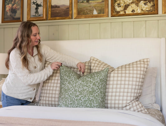 Woman displaying green and beige patterned cotton throw pillows on a bed with white headboard.