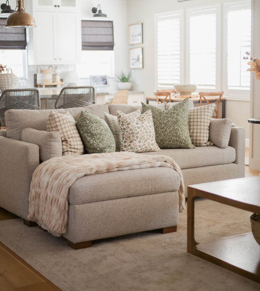 Beige upholstered sectional sofa with green and cream patterned pillows in bright living room setting.