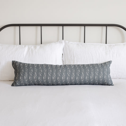 Sage green lumbar pillow with repeating leaf pattern displayed on bed with white pillows and black metal headboard.