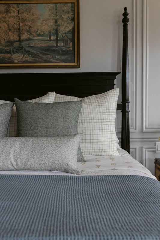 Grey and cream linen pillow with floral pattern displayed on bed with plaid and striped bedding.