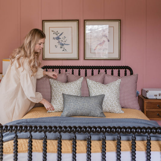 Woman in cream cardigan adjusting patterned pillows on bed with black tassel trim headboard against pink wall.