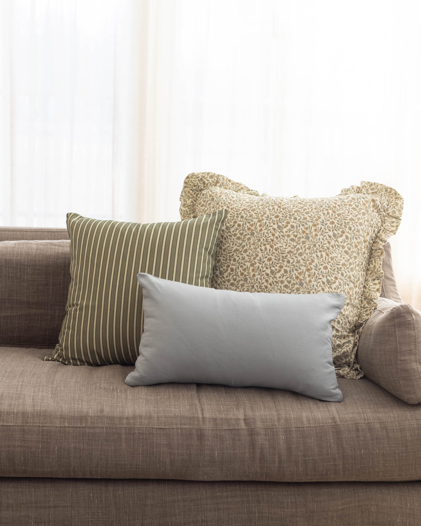 Light gray rectangular pillow with solid fabric displayed on beige sofa between striped and patterned accent pillows.