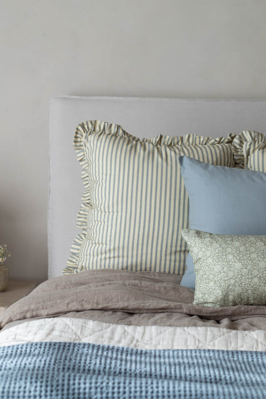 Green and white striped pillow with ruffled edges displayed on a bed with blue and patterned pillows.