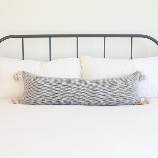 Cream tweed lumbar pillow with tassel corners displayed on a bed with white pillows.