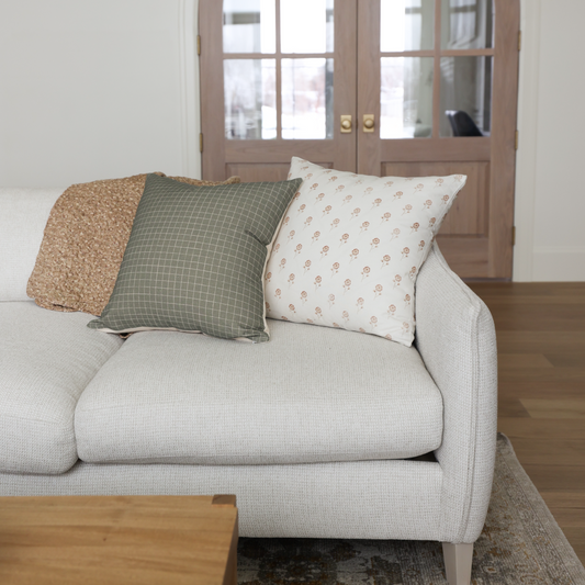 Cream upholstered armchair with rolled arms and three decorative pillows in tan, green grid, and cream floral patterns.