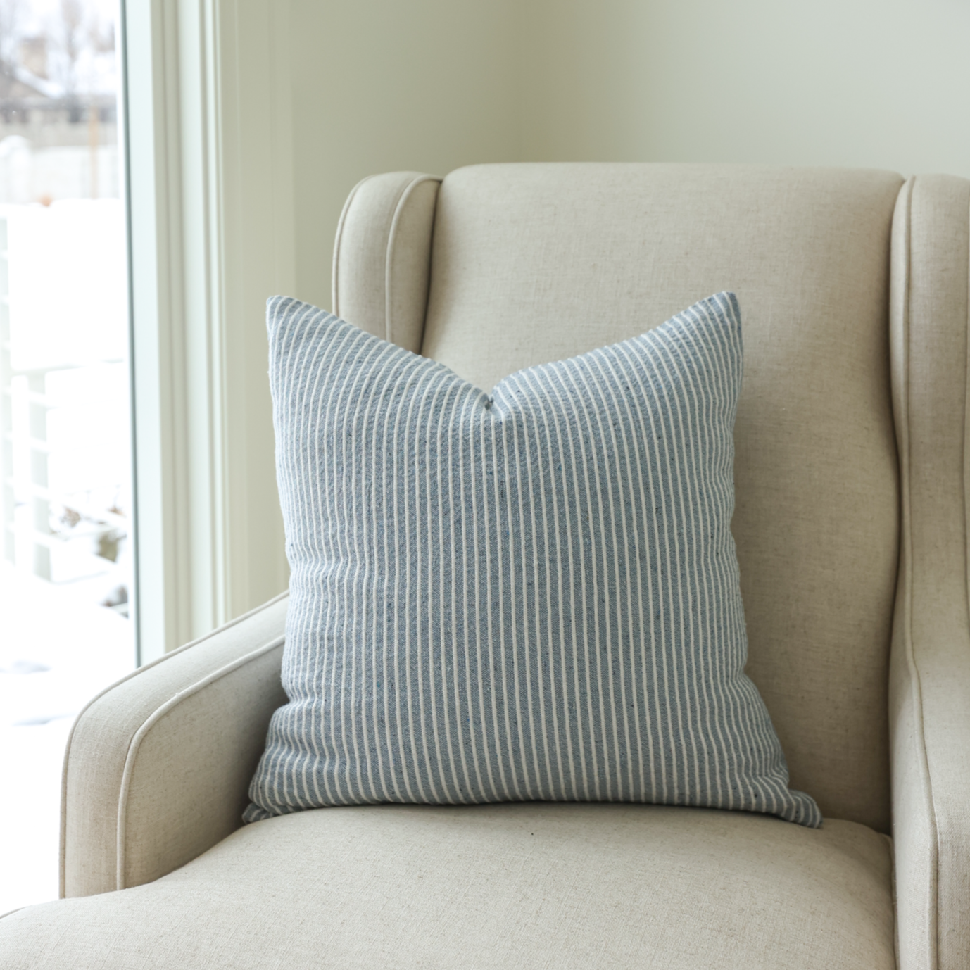 Blue and white striped pillow displayed on beige upholstered chair next to window.