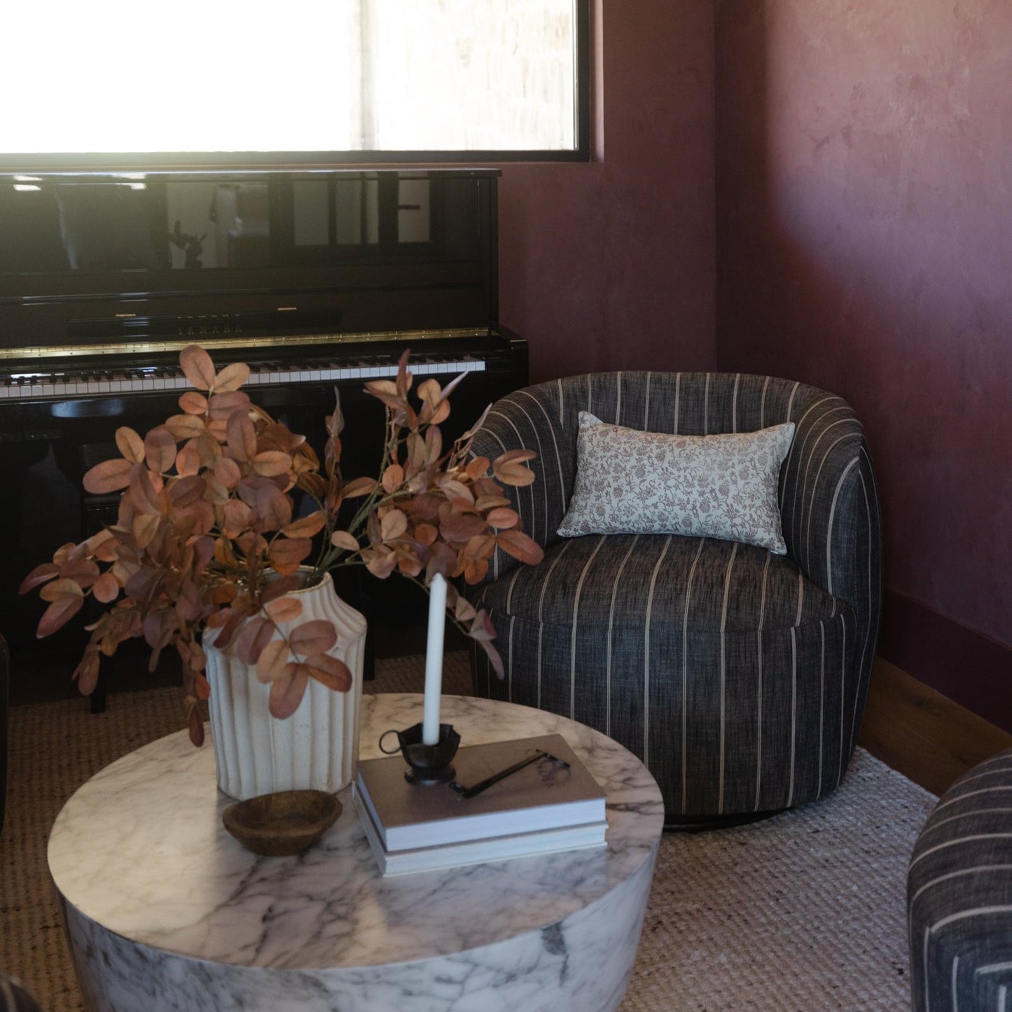 Burgundy wall with framed artwork above black piano, striped armchair with pillow, marble round table with dried flowers in v