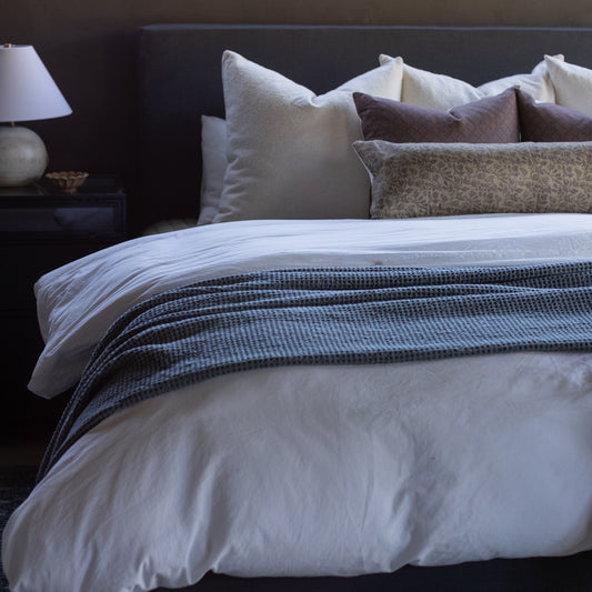 Faye pillow displayed on a bed with white bedding, navy striped throw, and coordinating cream and gray decorative pillows.