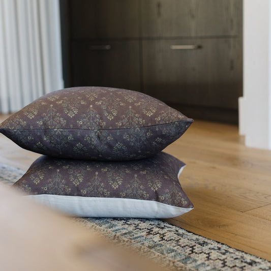 Stacked linen pillows in burgundy and navy with geometric damask pattern on wooden floor.