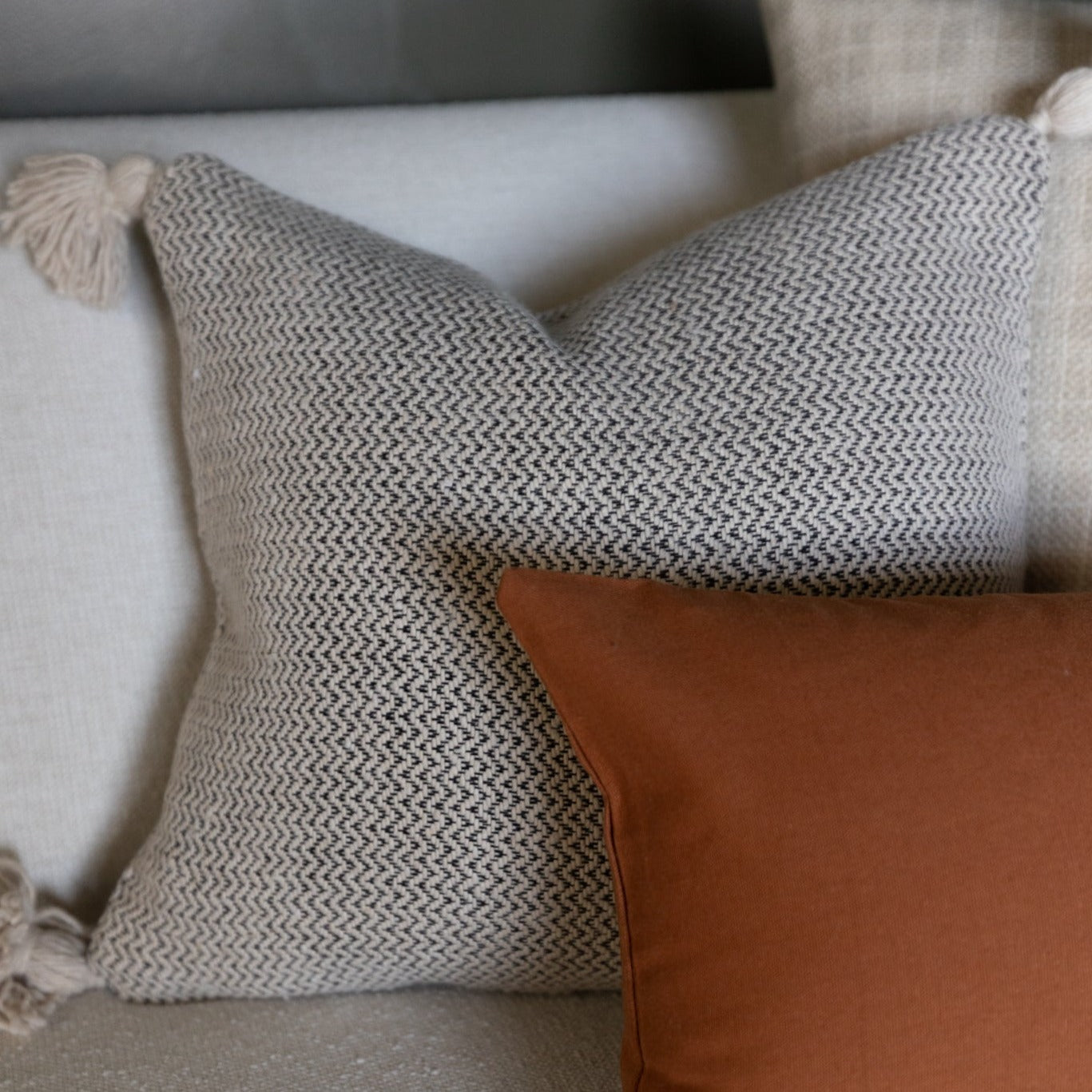 Cream-colored tweed pillow with herringbone pattern and corner tassel, displayed on furniture.