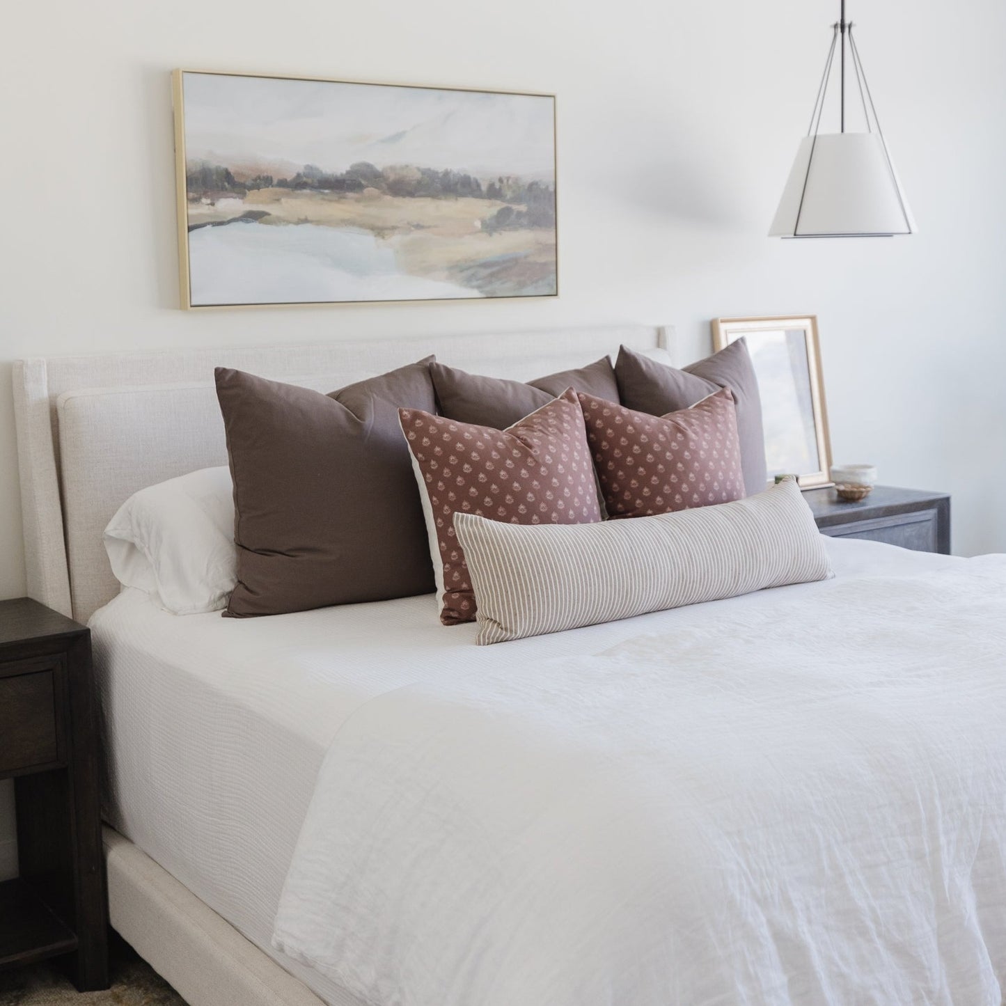 Bed with white linens and brown, mauve, and striped decorative pillows against a cream headboard.