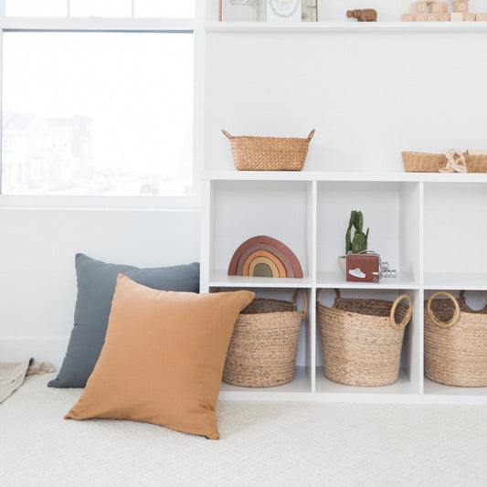 Terracotta-colored square pillow displayed on white shelving with woven baskets and decor.