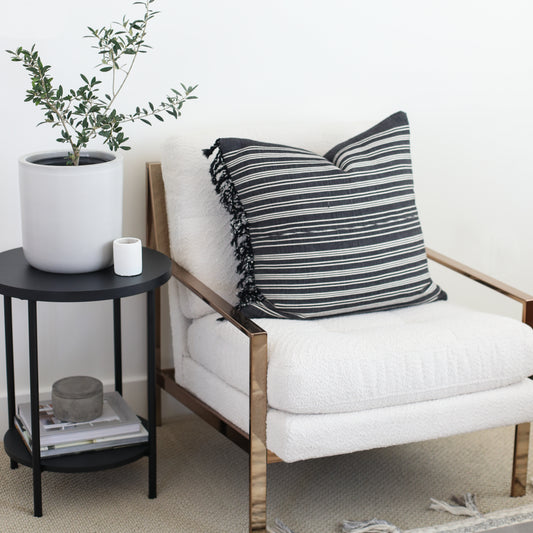 Black and white striped pillow with fringe trim on natural wood-frame chair with cream cushion.