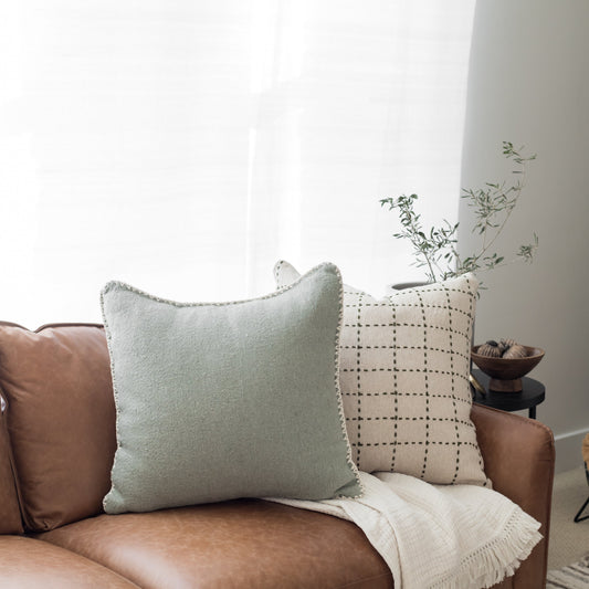 Light green textured pillow with frayed edges displayed on a brown leather chair.