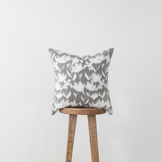 Gray and white floral patterned pillow with white flowers and green leaves on wooden stool.