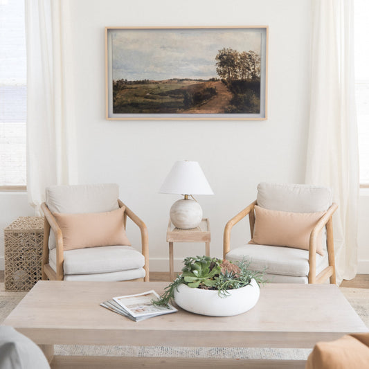 Framed landscape painting with natural wood frame hanging above two upholstered chairs in neutral tones.