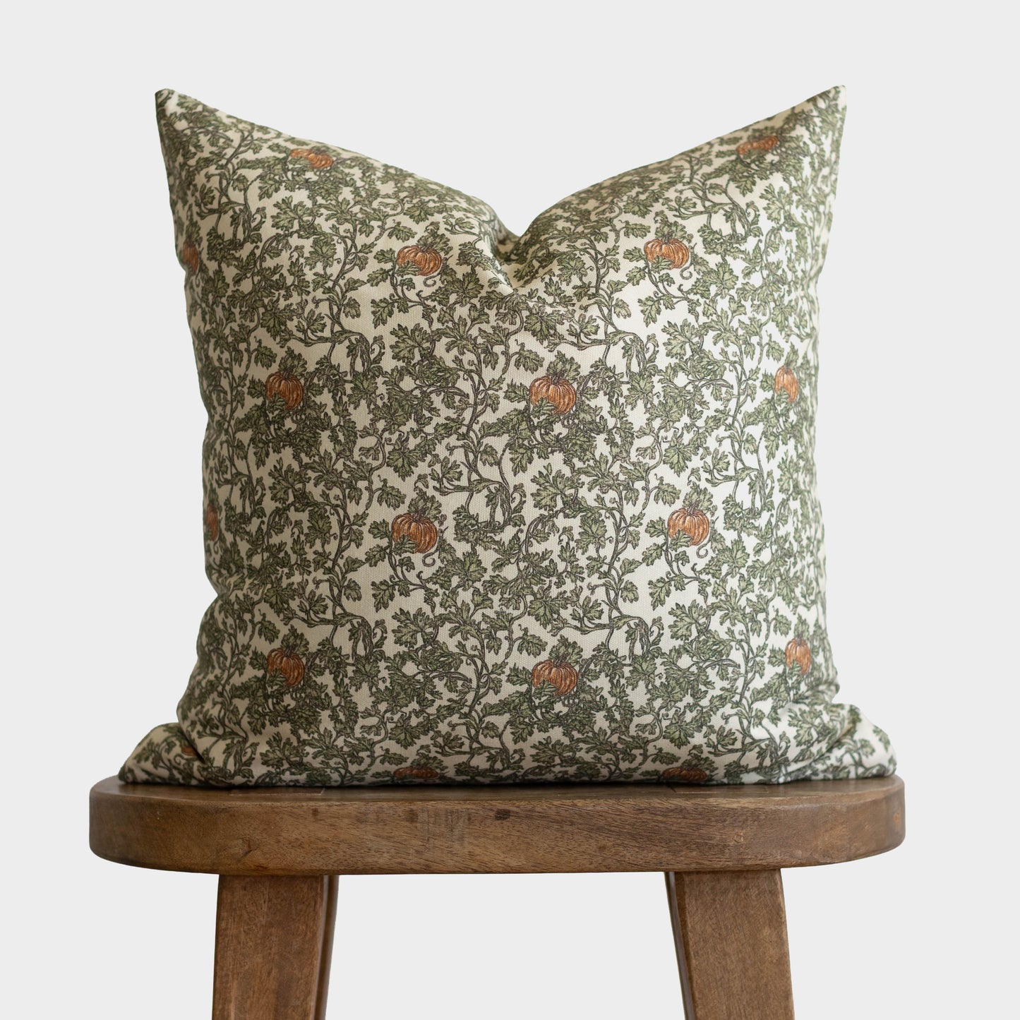 Cream linen pillow with olive green foliage and rust-colored gourds pattern displayed on wooden stool.