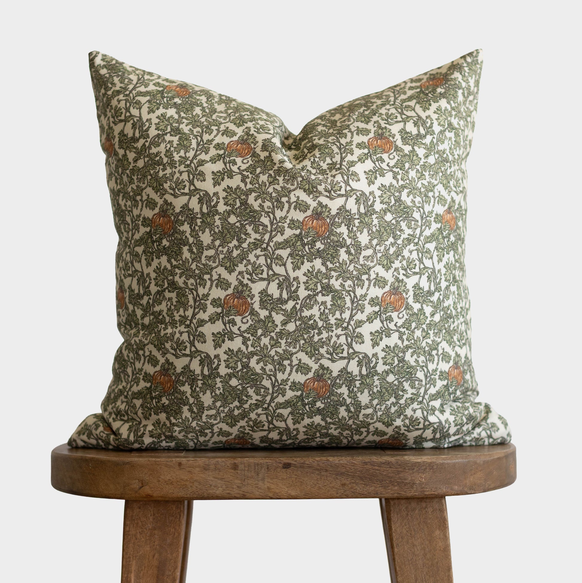 Cream linen pillow with olive green foliage and rust-colored gourds pattern displayed on wooden stool.