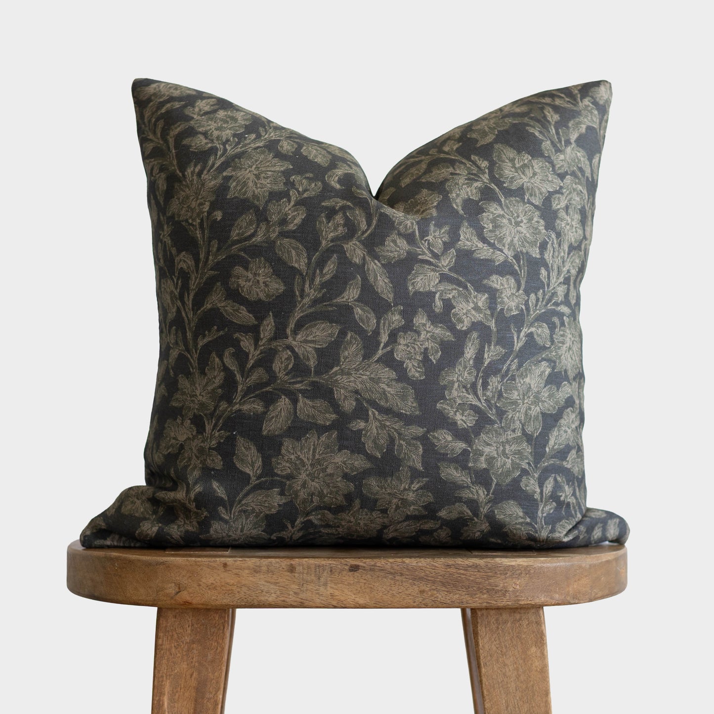 Dark charcoal pillow with gold botanical leaf pattern, displayed on wooden stool.