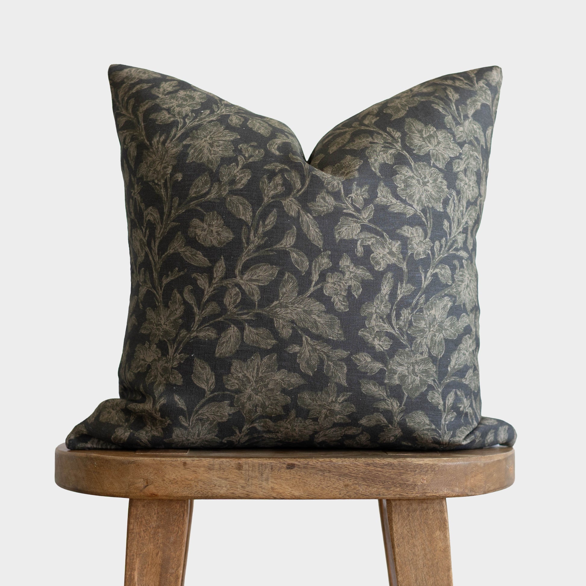 Dark charcoal pillow with gold botanical leaf pattern, displayed on wooden stool.