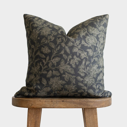 Dark charcoal pillow with gold botanical leaf pattern, displayed on wooden stool.