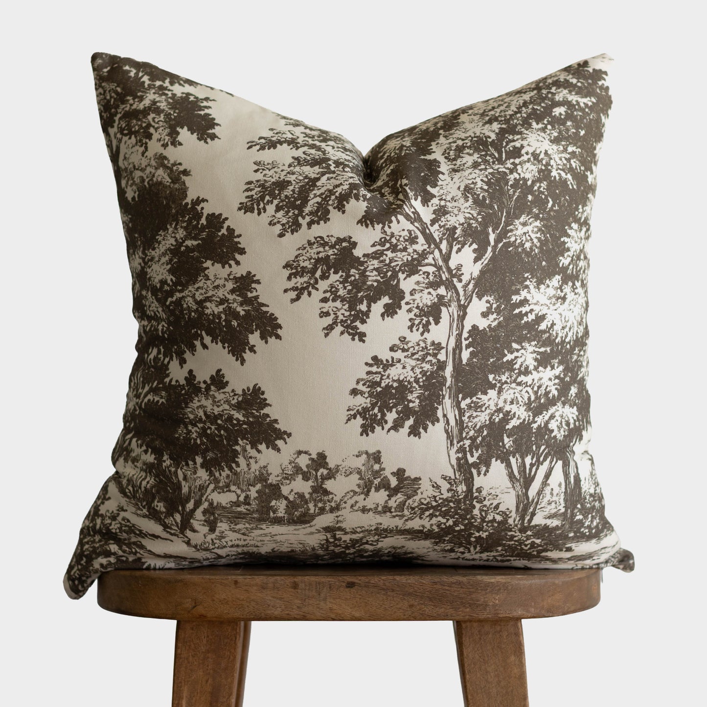 Cream pillow with dark brown toile forest and tree landscape pattern, displayed on wooden stool.