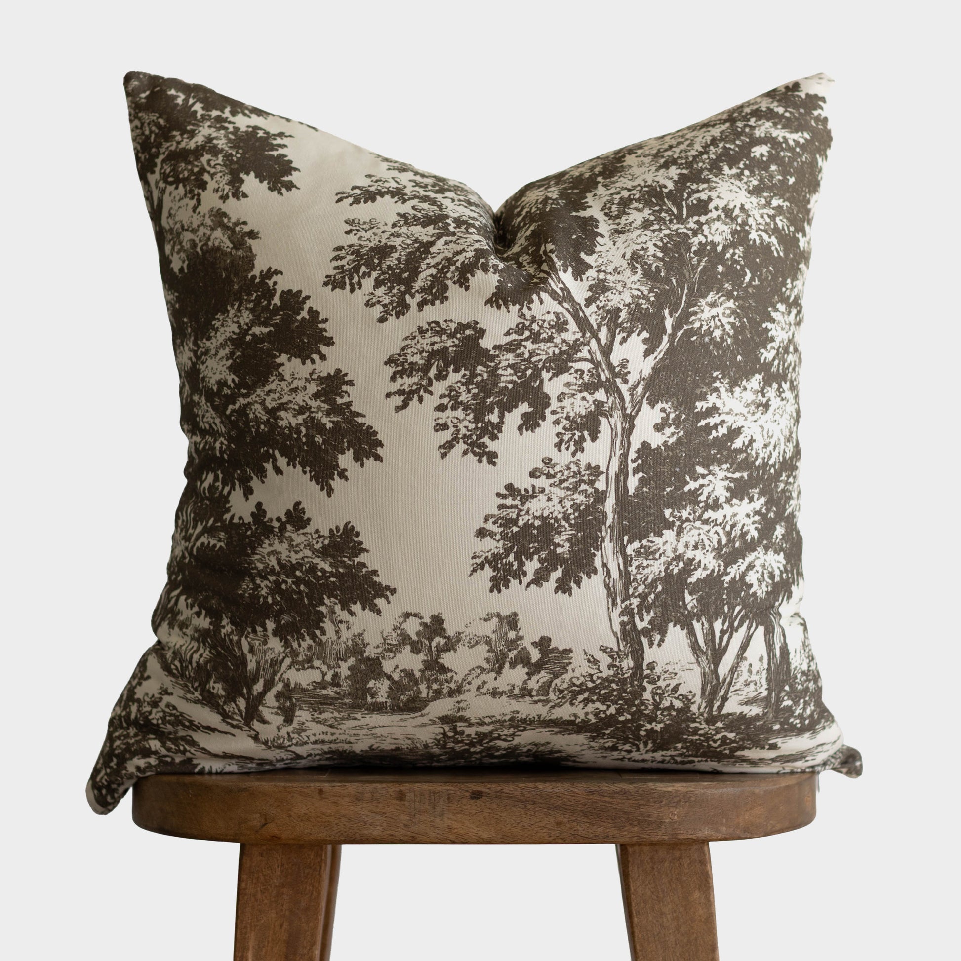Cream pillow with dark brown toile forest and tree landscape pattern, displayed on wooden stool.