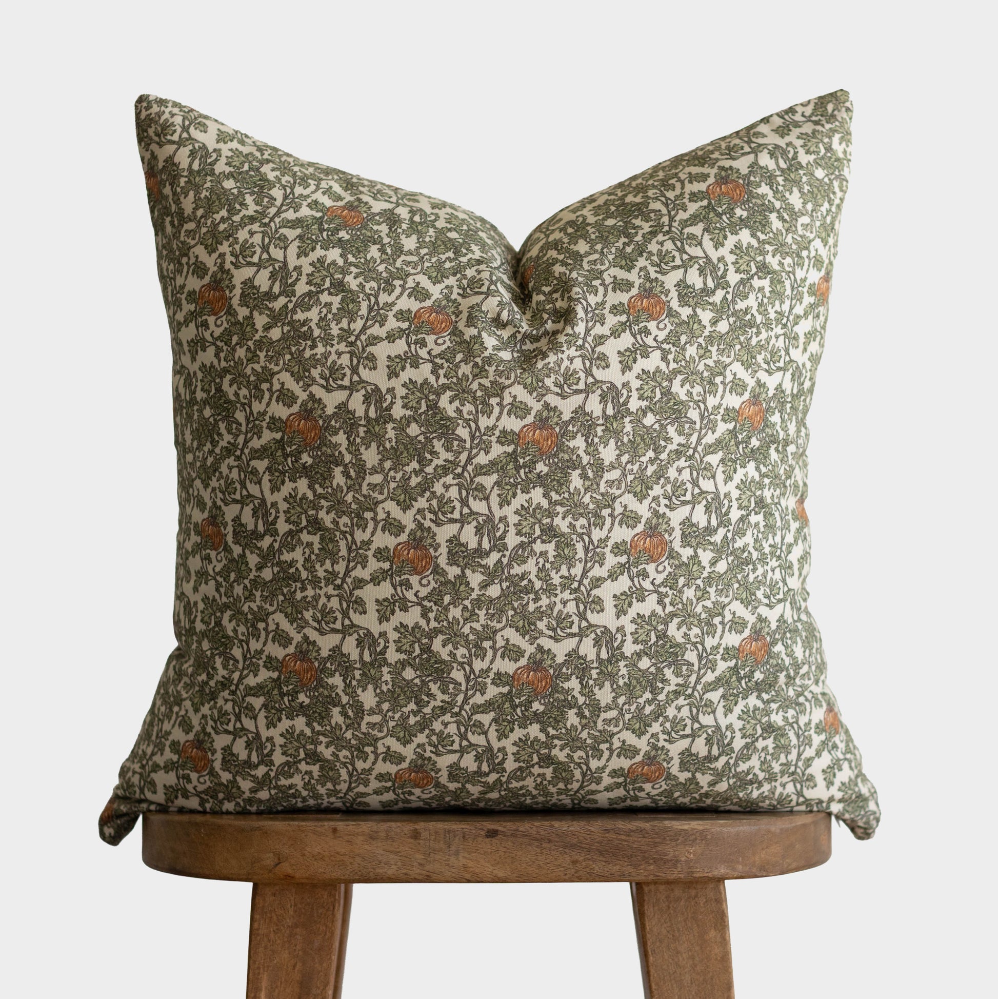 Cream pillow with green botanical print and rust-colored gourds, displayed on wooden stool.