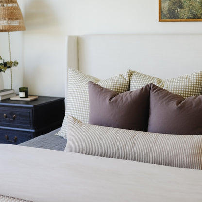 Tan lumbar pillow displayed on a bed with checkered and striped pillows in a neutral bedroom setting.