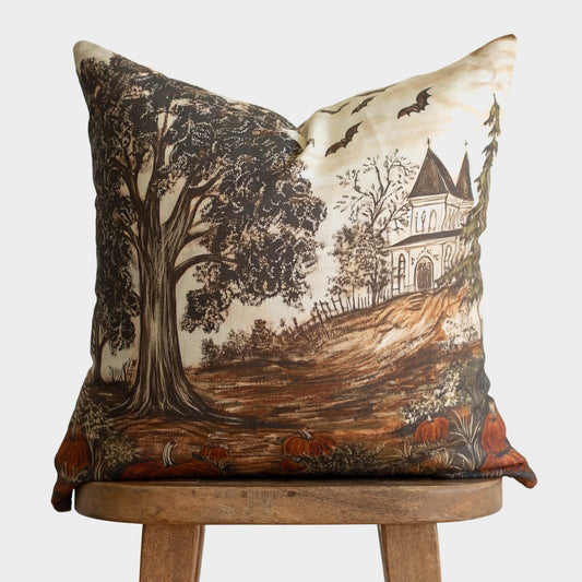 Linen pillow with illustration of gnarled tree, haunted house, bats, pumpkins, and bare branches in brown and cream tones.