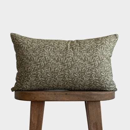 Green and tan leaf patterned pillow displayed on wooden stool against neutral background.