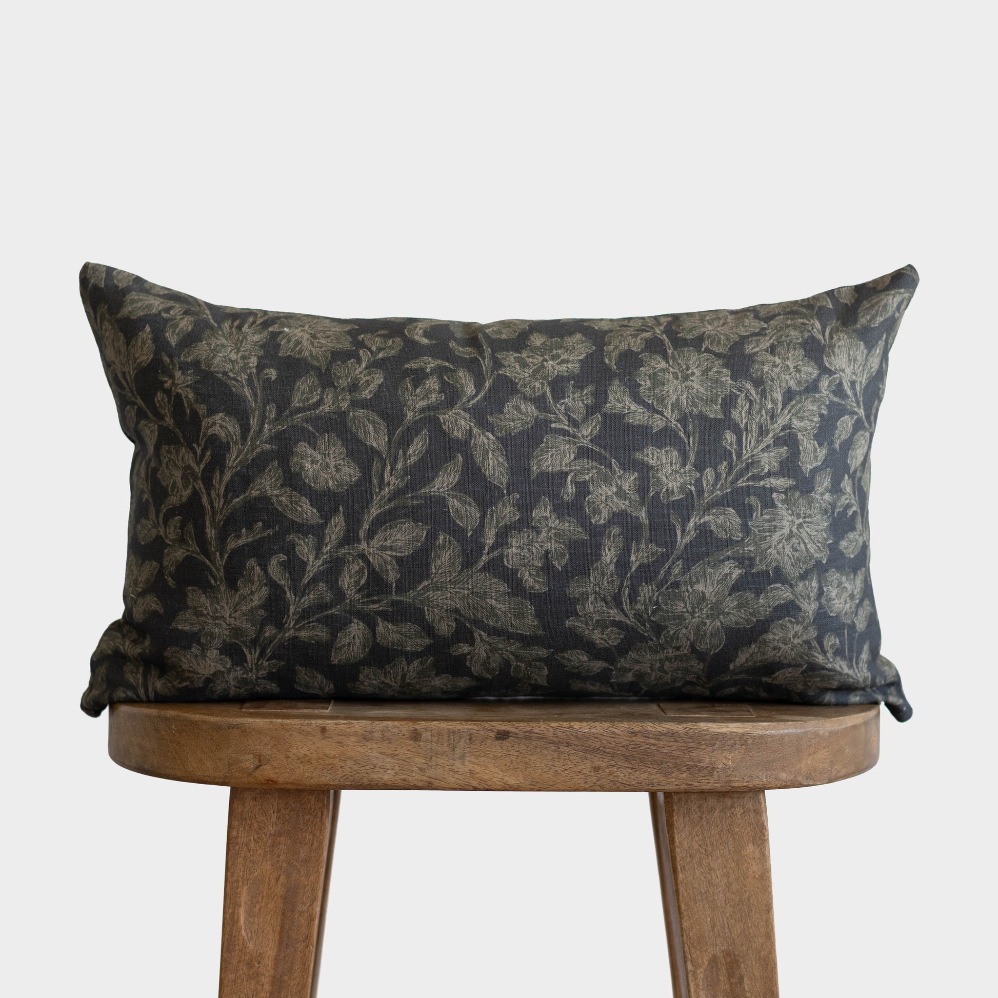 Charcoal pillow with gold botanical print placed on wooden stool.
