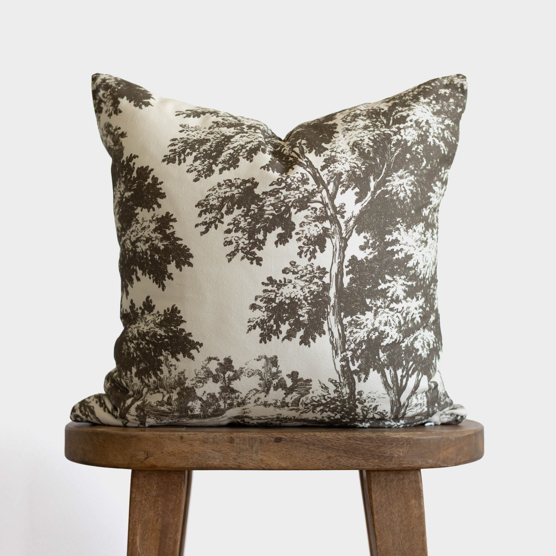 Cream and brown toile patterned throw pillow with tree and foliage design, displayed on wooden stool.